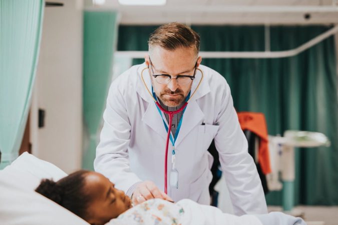 Un médecin en urgence portant des lunettes utilise un stéthoscope pour examiner attentivement une jeune patiente allongée dans un lit d'hôpital.
