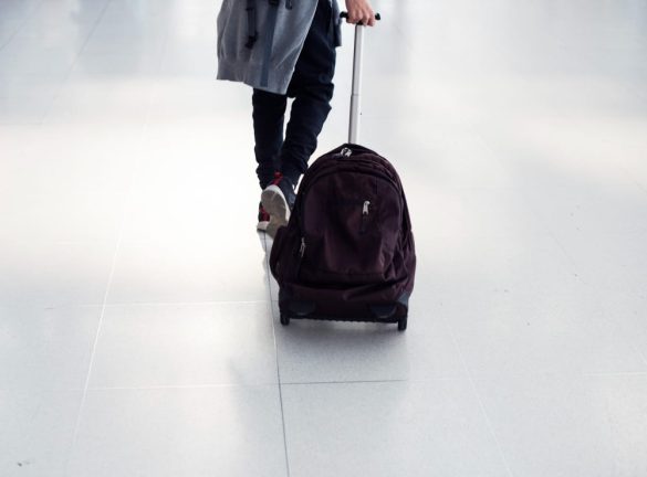 Une personne vue de dos tire un grand cartable à roulettes noir avec une poignée télescopique dans un terminal d'aéroport au sol carrelé