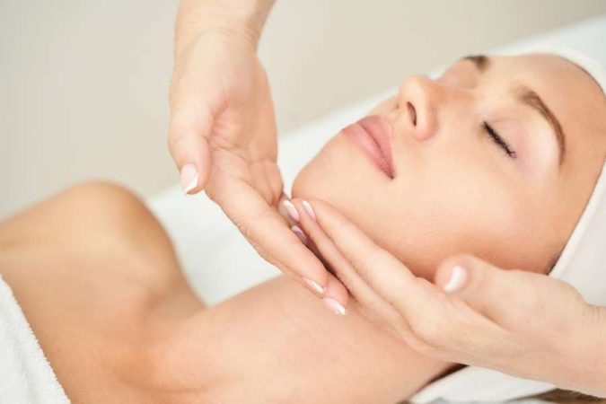 Gros plan des mains d'une esthéticienne effectuant un massage du visage sur une cliente détendue, illustrant la douceur du layering.