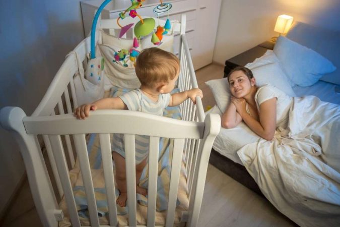 Bébé se réveille la nuit : un tout-petit debout dans son lit à barreaux regarde sa maman.