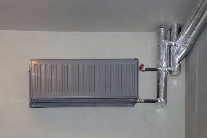 Un radiateur en fonte d'aluminium gris suspendu sur un mur clair, raccordé à des tuyaux calorifugés.