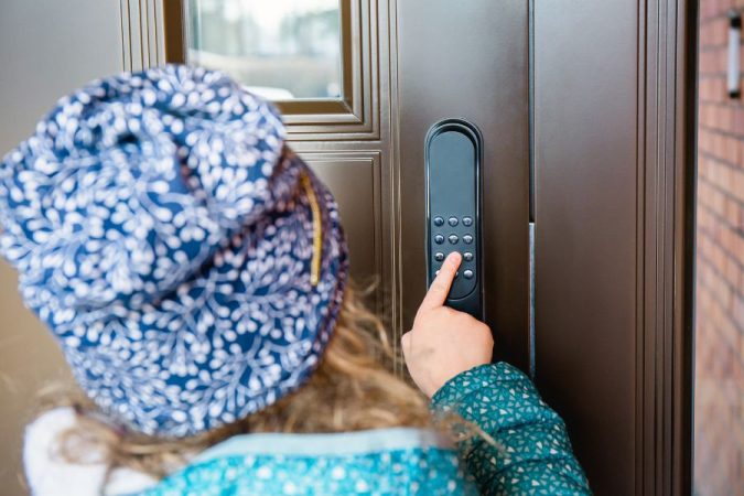 Une personne de dos avec un bonnet à motifs appuie sur les touches d'une serrure à code mécanique noire montée sur une porte d'entrée marron.